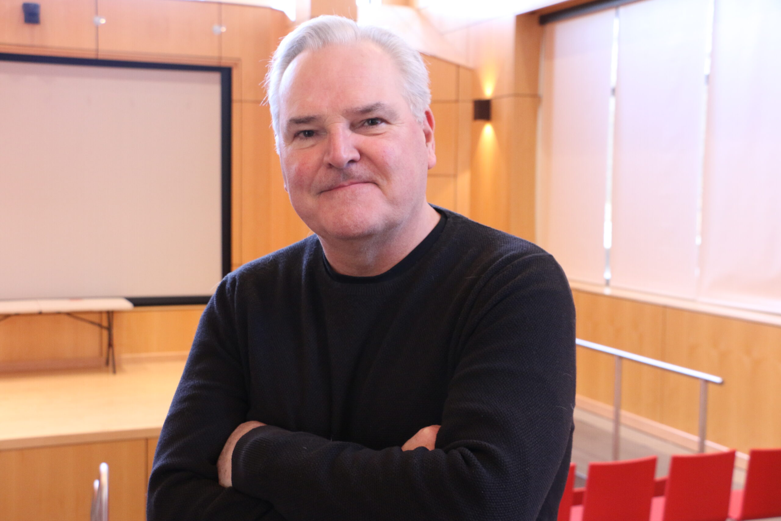Jody Wood wears a dark, long sleeved shirt, and a pleasant expression, standing in a brightly lit presentation space.