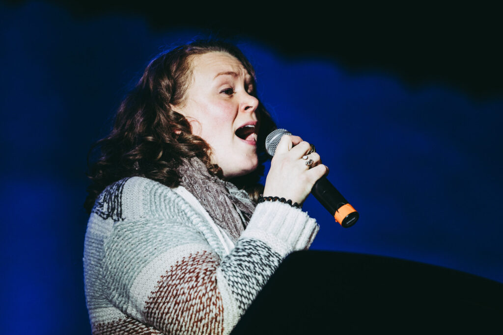 Stephanie Overton-Hall is wearing a woven striped cardigan and her hair is brown and curly. She holds a microphone and sings passionately against a dark black background to her invisible audience.