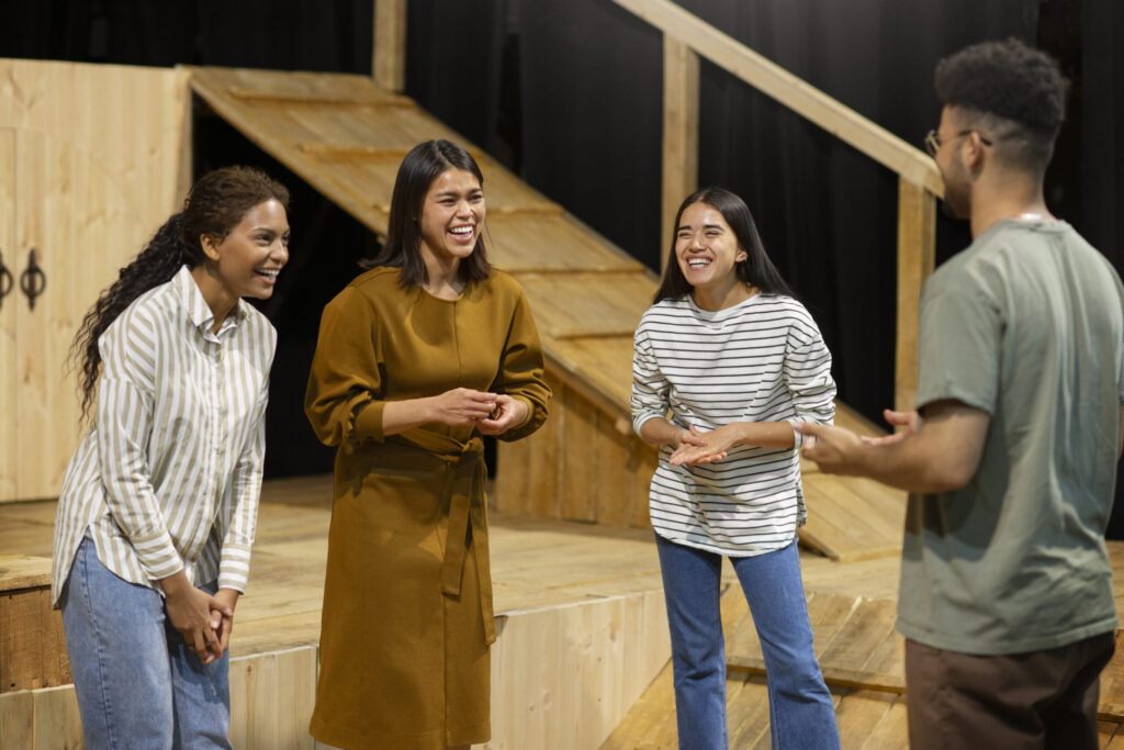 Four actors of varied ethnicities stand on a light wood stage, laughing naturally together in their scene. A light wood ramp stands behind them in the background, backed by a large black stage curtain.