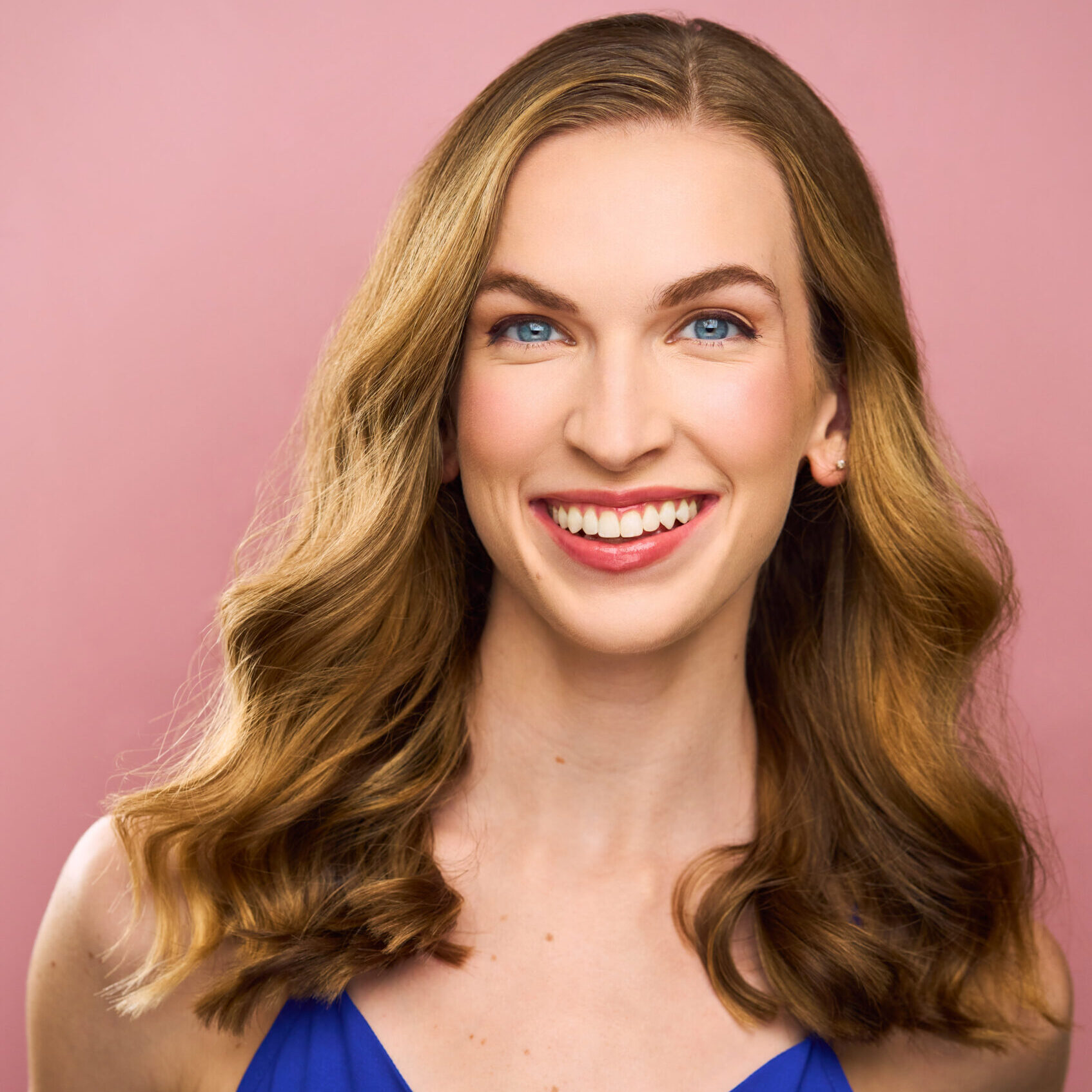 Headshot of Allison Reed, white female dancer with a bright, friendly smile, cool blue eyes, and long wavy blonde hair, wearing a dark blue tank top in front of a solid blush pink background.