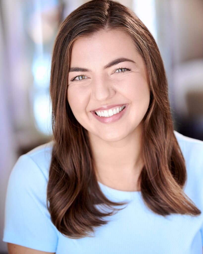 Headshot of Amalea Hammitt, white female dancer with a bright, friendly smile, hazel eyes, and long straight brown hair, wearing a light blue tee in front of a blurred background.