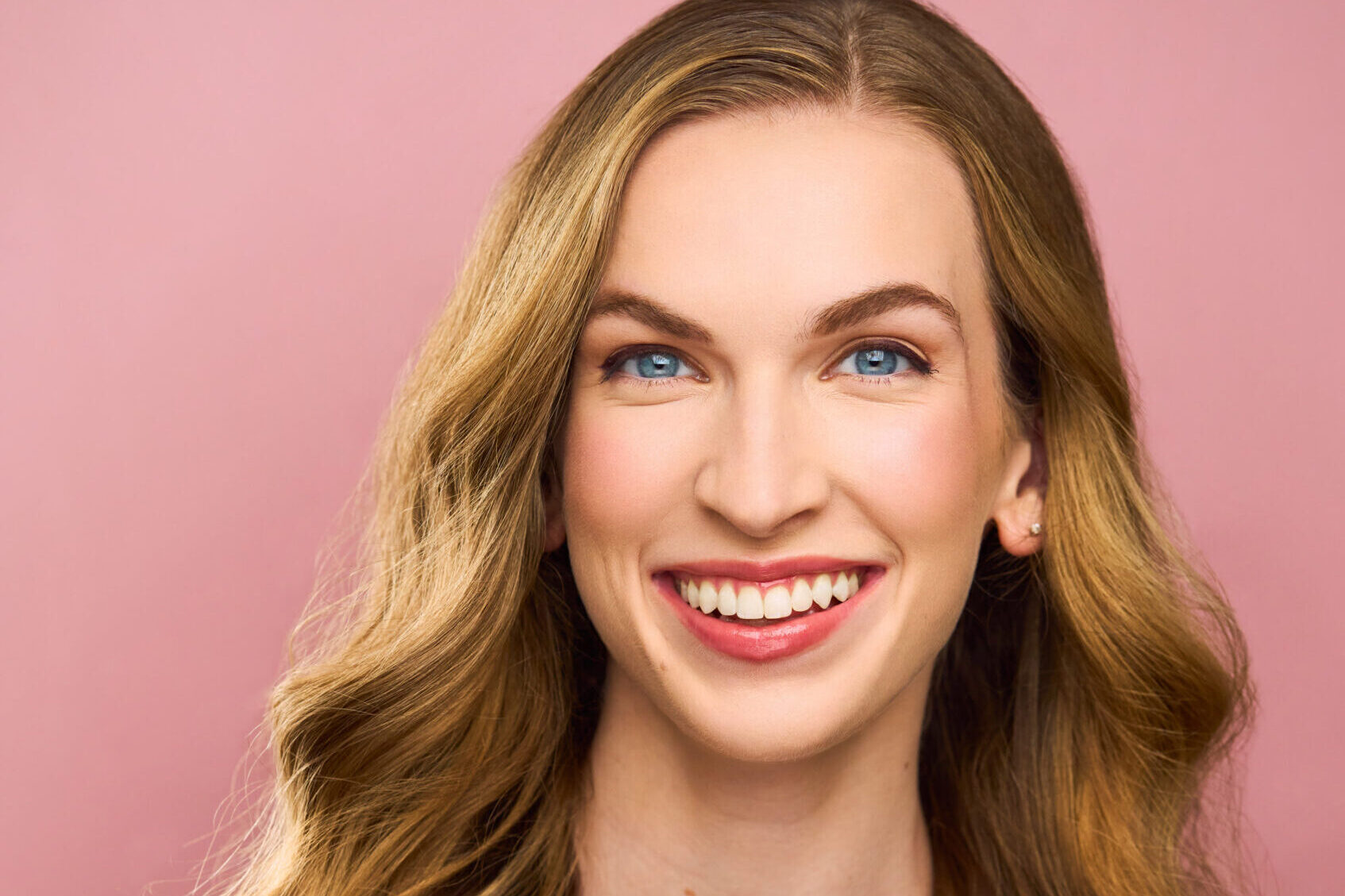 Headshot of Allison Reed, white female dancer with a bright, friendly smile, cool blue eyes, and long wavy blonde hair, wearing a dark blue tank top in front of a solid blush pink background.