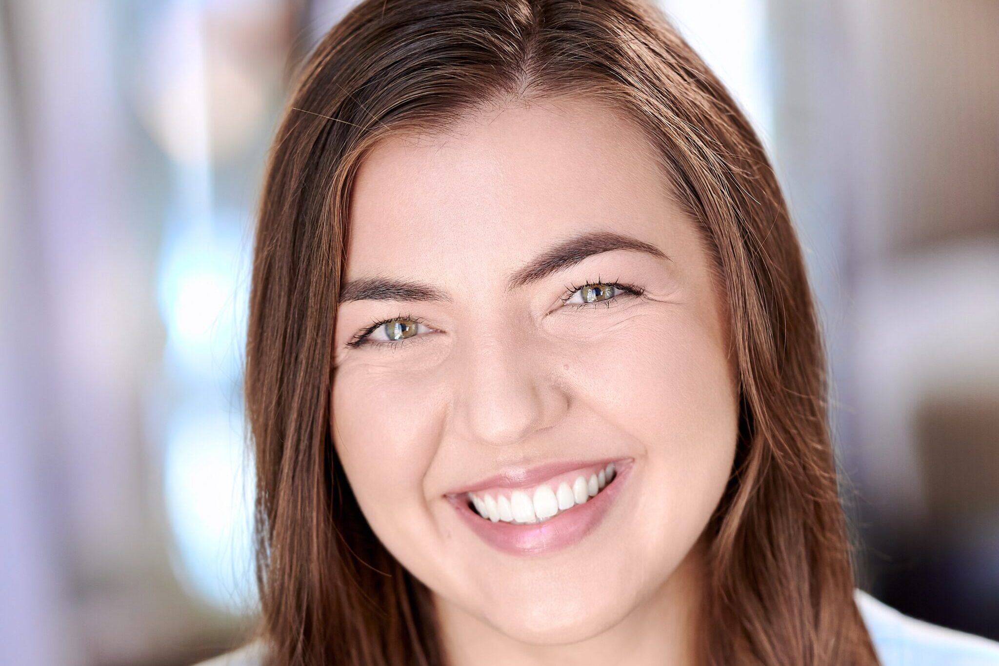 Headshot of Amalea Hammitt, white female dancer with a bright, friendly smile, hazel eyes, and long straight brown hair, wearing a light blue tee in front of a blurred background.