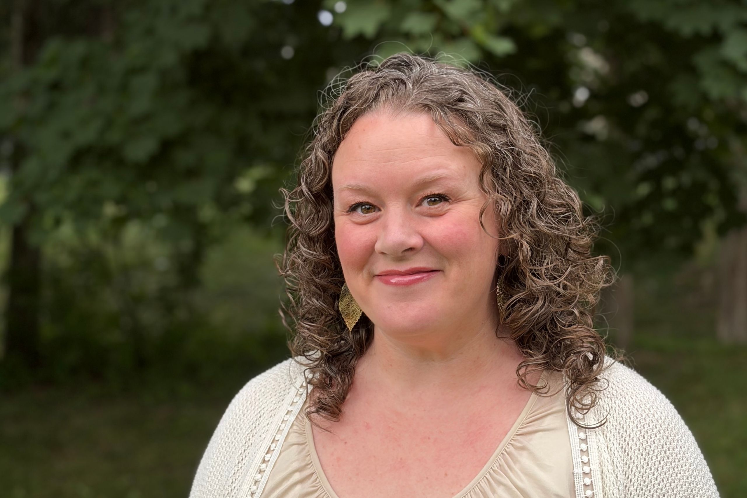 Stephanie Overton-Hall, white female with brown curly hair and brown eyes, with a pleasant, close-lipped smile. She is wearing a beige shimmery shirt, an off-white cardigan, and dangling gold earrings that look like almond shaped leaves. Background is a slightly blurred natural setting with greenery.